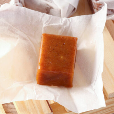 Homemade buttery dark brown sugar caramels wrapped in white paper, presented on a wooden tray with a checkered cloth in the background, on a wooden surface. There's an unwrapped caramel in focus at the front of the image.