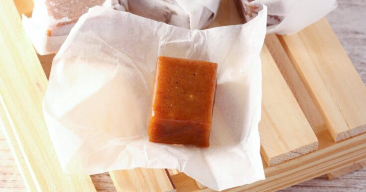 Homemade buttery dark brown sugar caramels wrapped in white paper, presented on a wooden tray with a checkered cloth in the background, on a wooden surface. There's an unwrapped caramel in focus at the front of the image.