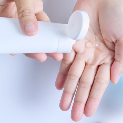 Two hands are shown, one dispensing a clear homemade diy hair gel from a white, cylindrical container onto the open palm of the other hand. The background is plain and minimal, emphasizing the hands and the container—perfect for showcasing your homemade organic hair gel recipe