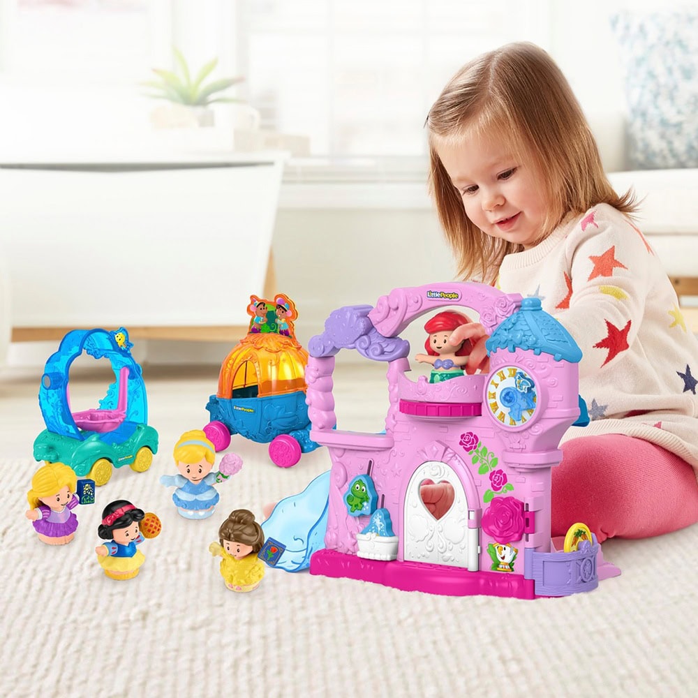 A young child plays with a pink and blue dollhouse with a heart-shaped door, surrounded by colorful toy figures and a mini carriage. The scene resembles a whimsical Play Set, and the child is sitting on a light carpeted floor in a well-lit room. A cushioned seat is visible in the background.