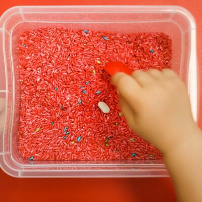 Child's hands engaging in sensory play with a scoop and red dyed rice decorated with small multicolored objects in a clear plastic bin on a red background for a Valentine's Day sensory tub.