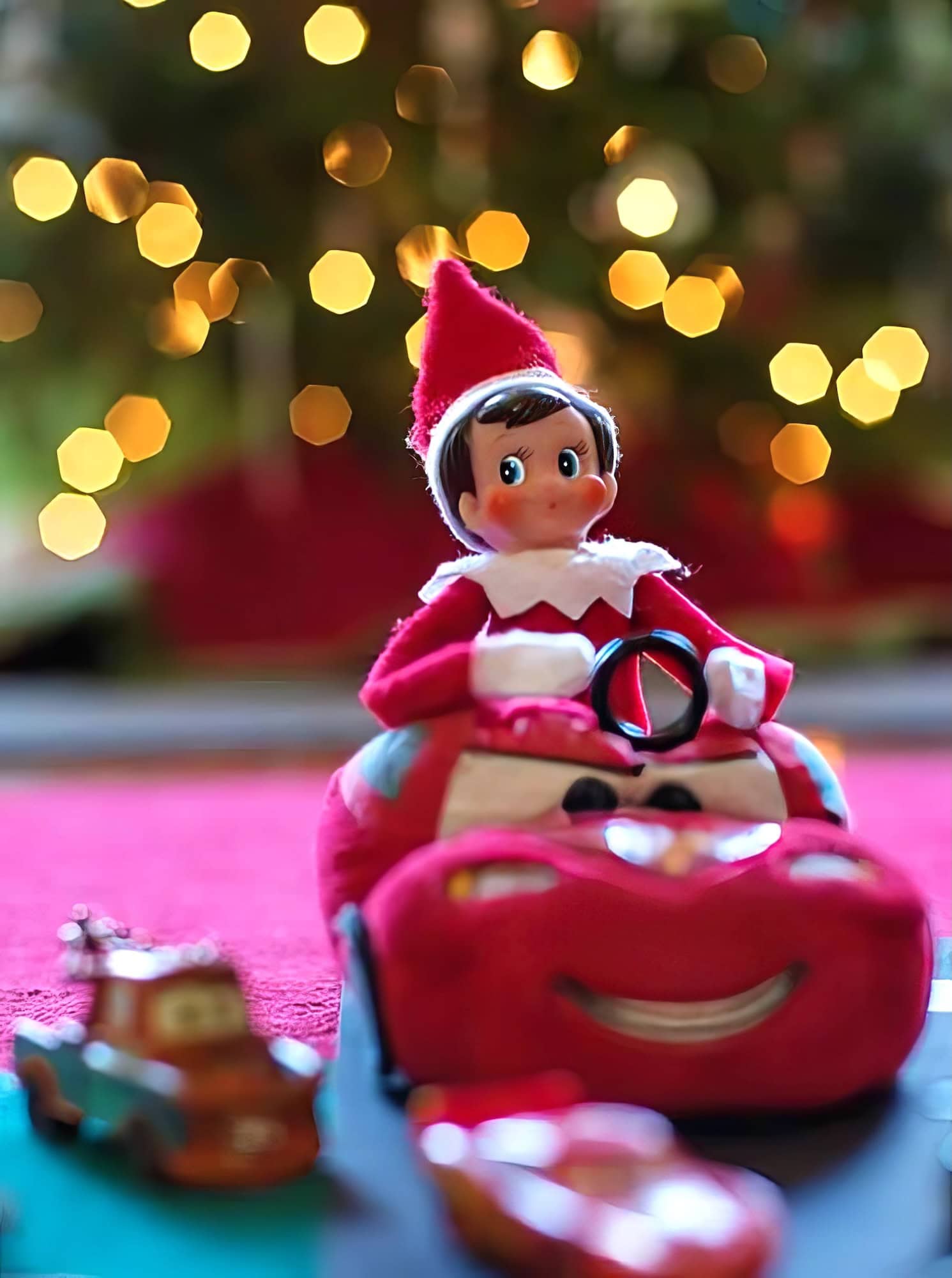 A festive Elf on the Shelf toy, inspired by Disney Elf on the Shelf ideas, sits in a plush red car with a steering wheel. Behind it, a Christmas tree glows warmly with blurred lights. Two small toy cars are parked in the foreground on a vibrant red surface.