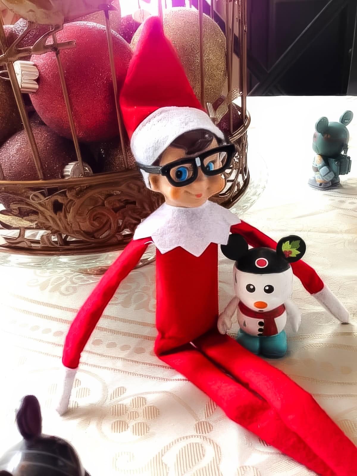 An Elf on the Shelf doll with glasses and a red outfit sits on a table, holding a small decorative snowman wearing Mickey Mouse ears. This charming Disney Elf on the Shelf idea is completed with a basket of large red and gold ornaments in the background.