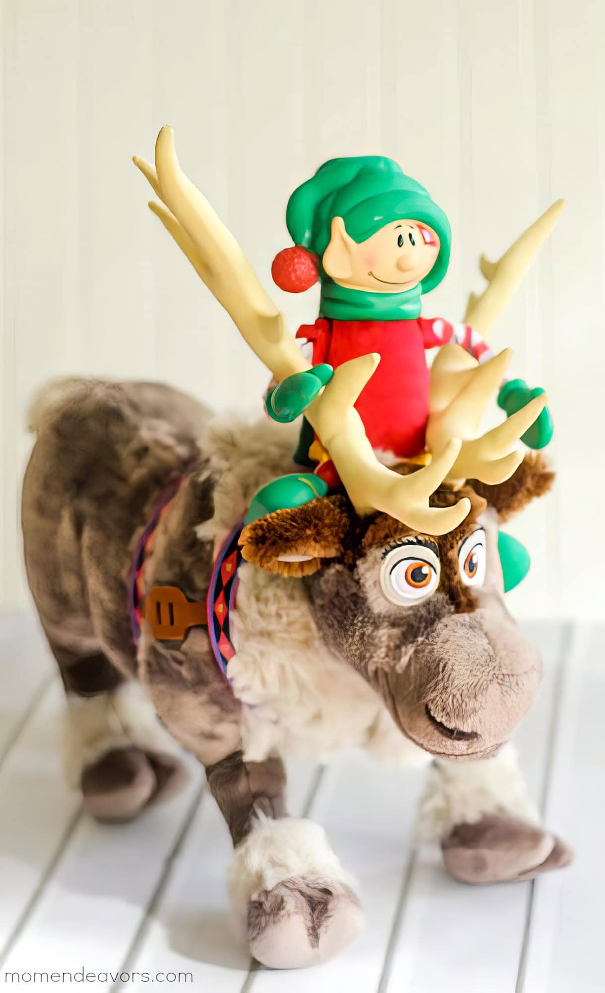 A plush reindeer toy with large antlers, Sven from Frozen, stands on a white surface. Nestled between the antlers is a small Elf on the Shelf figure in a green hat and red outfit, reminiscent of Disney elf on the shelf ideas. The reindeer has a cheerful expression, adding charm to this whimsical scene.