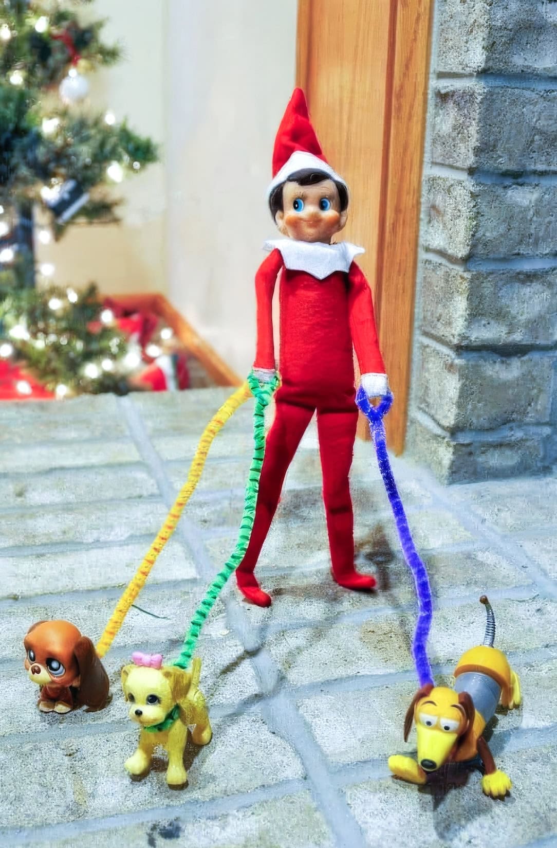 A Disney elf on the shelf idea comes to life with a toy elf dressed in red, standing by the Christmas tree. It holds colorful pipe cleaners as leashes for three uniquely featured toy dogs, while festive holiday décor lights up the background.