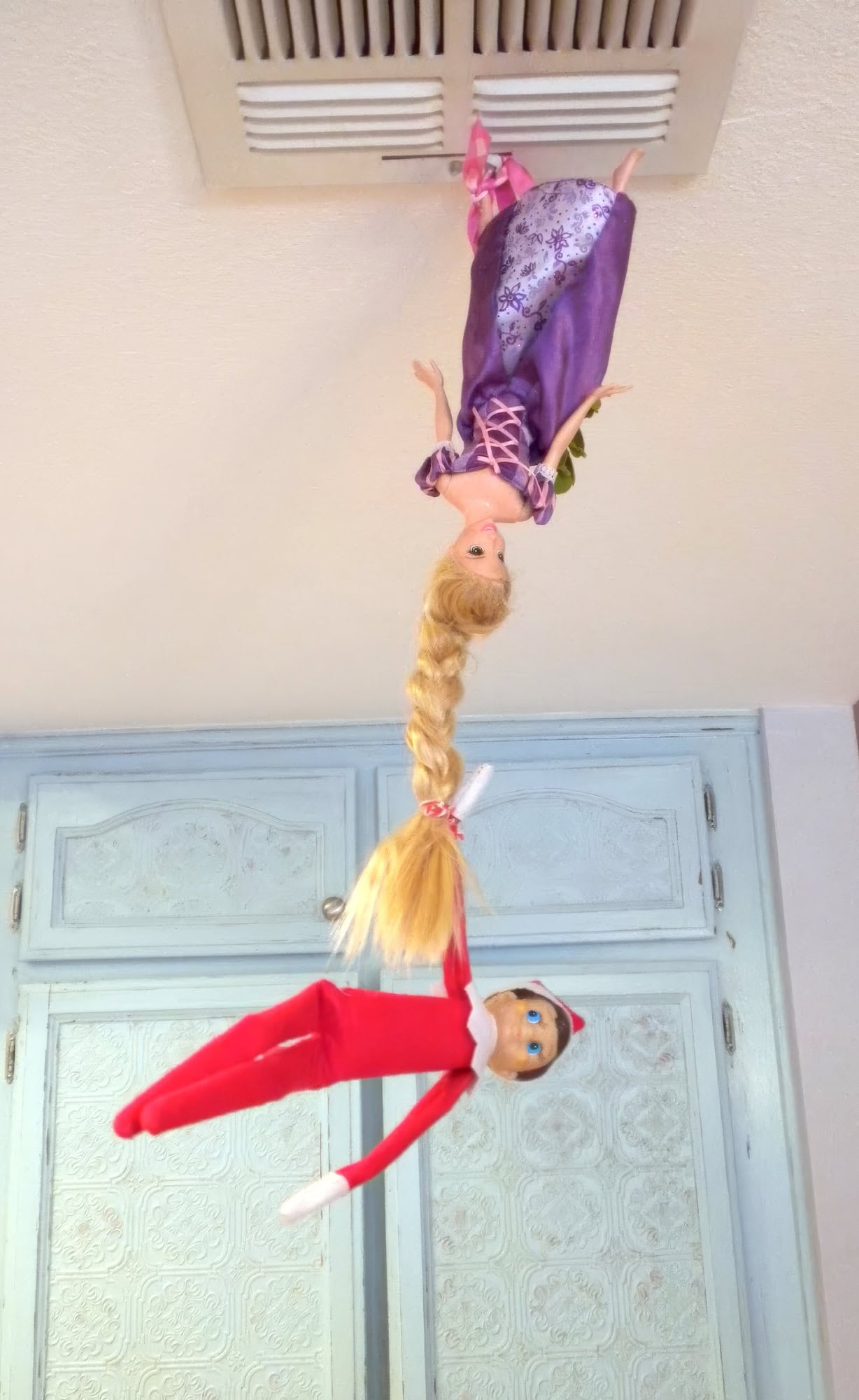 A Disney-inspired scene unfolds as a doll with long blonde hair, clad in a purple gown, hangs upside down from a ceiling vent. Below her, an elf on the shelf in a red outfit dangles playfully, clutching the doll's hair. Blue cabinets provide the whimsical backdrop to this imaginative setup.