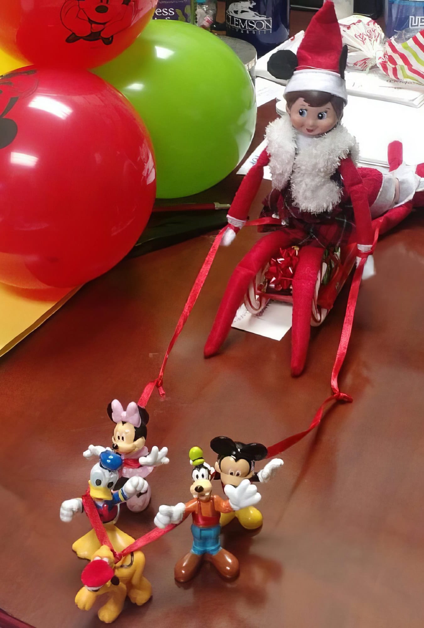 A festive Disney scene unfolds on the table with a charming elf on the shelf dressed in red, perched on a sled. This whimsical elf is "pulled" by toy figures—Minnie, Mickey, Goofy, and Pluto—with red ribbons as reins. Bright balloons provide a joyful backdrop to this delightful holiday setup.