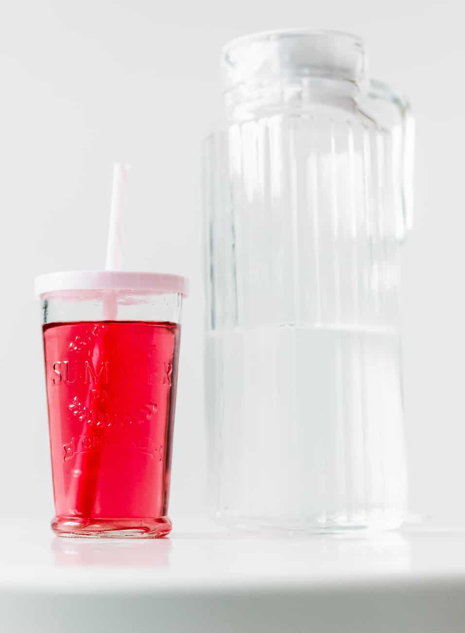 clear red drink in a clear glass glass with a white lid and straw next to a pitcher with water.
