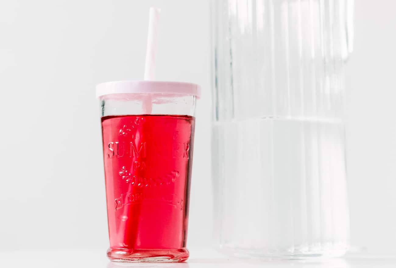 Clear pink beverage in clear glass with white straw and lid next to a pitcher