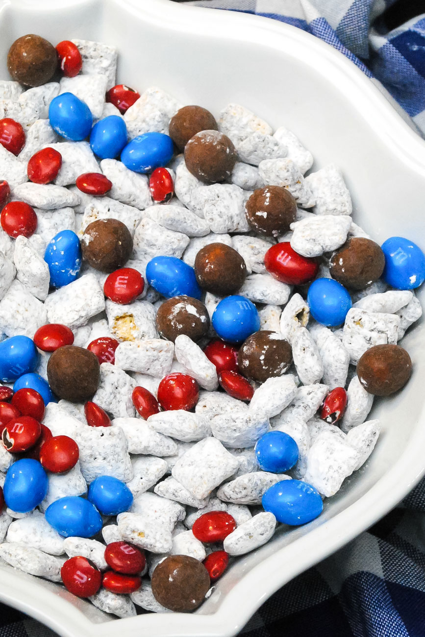 red, white and blue dessert snack mix with 4th of July candy and coated cereal in a white bowl over a blue and white napkin.