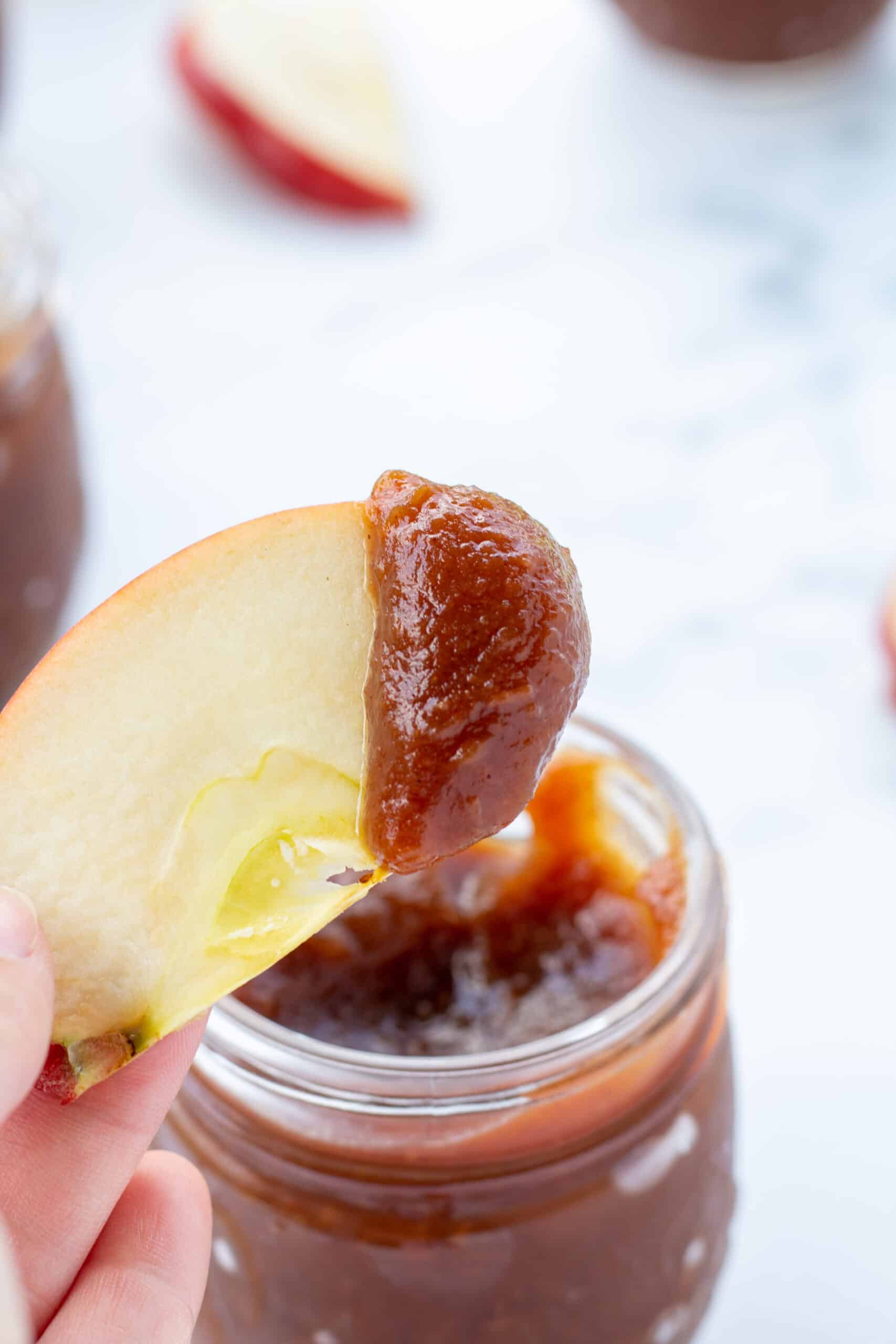 an apple slice dipped in homemade apple butter held by a hand over a glass jar of canned apple butter