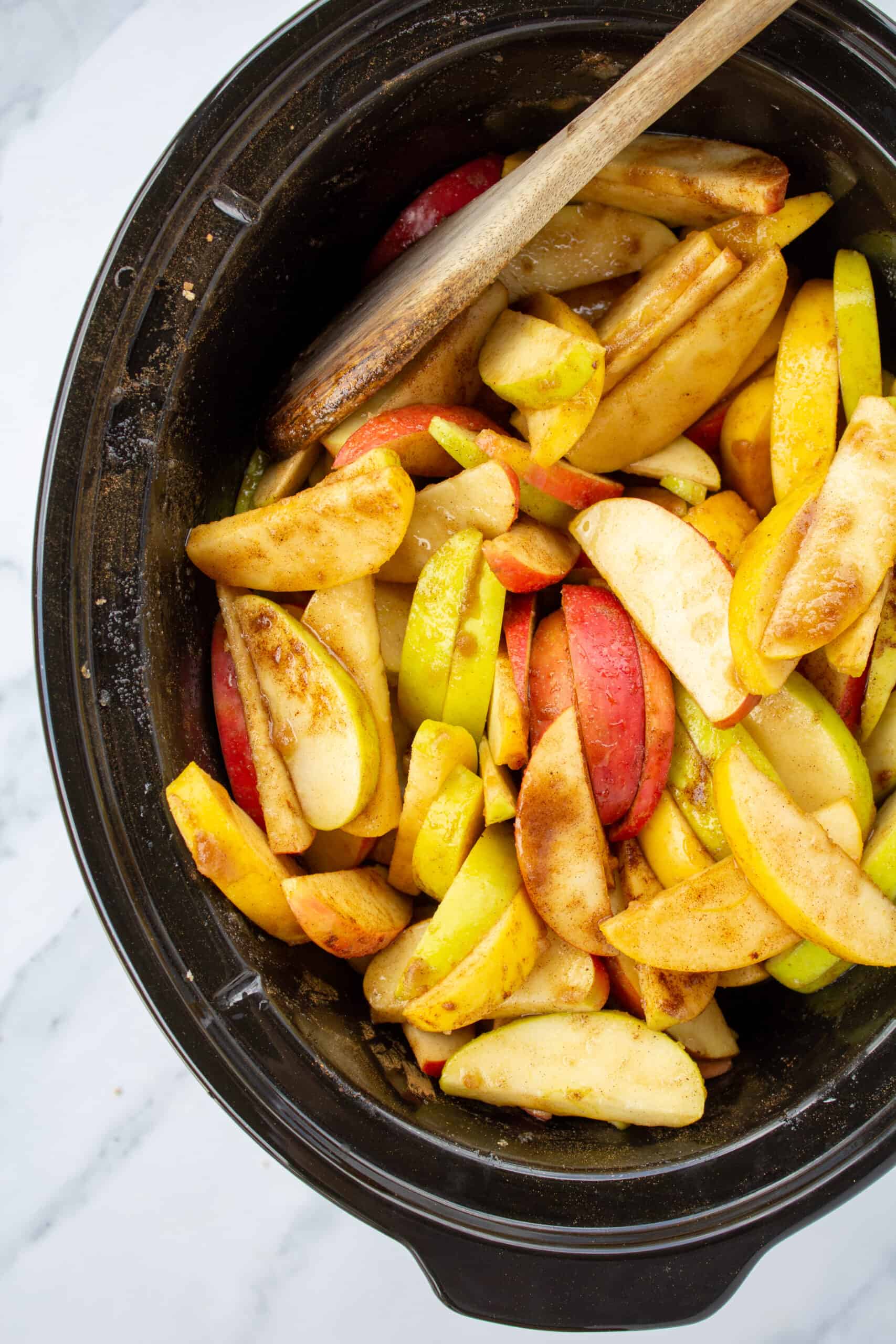 crockpot full of coated apples and a wooden spoon sitting on a marble counter