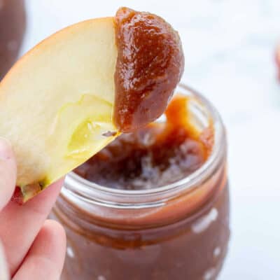 how to make apple butter with a hand holding an apple slice dipping in jar of apple preserves