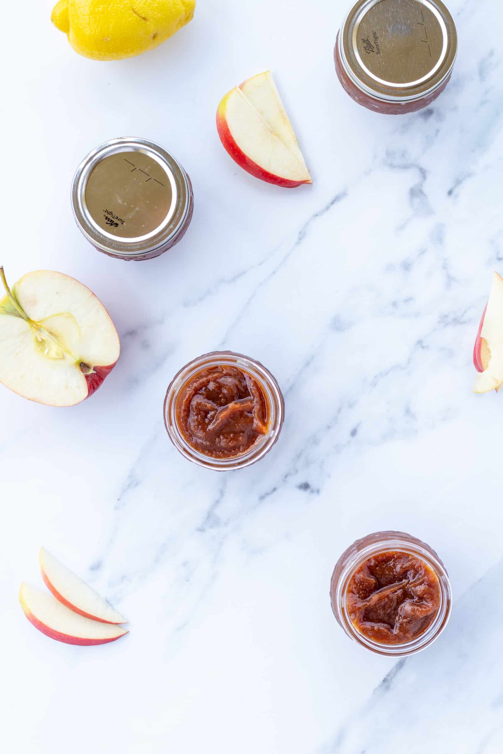 jars of apple butter, slices of apples, and a lemon sitting on a white counter