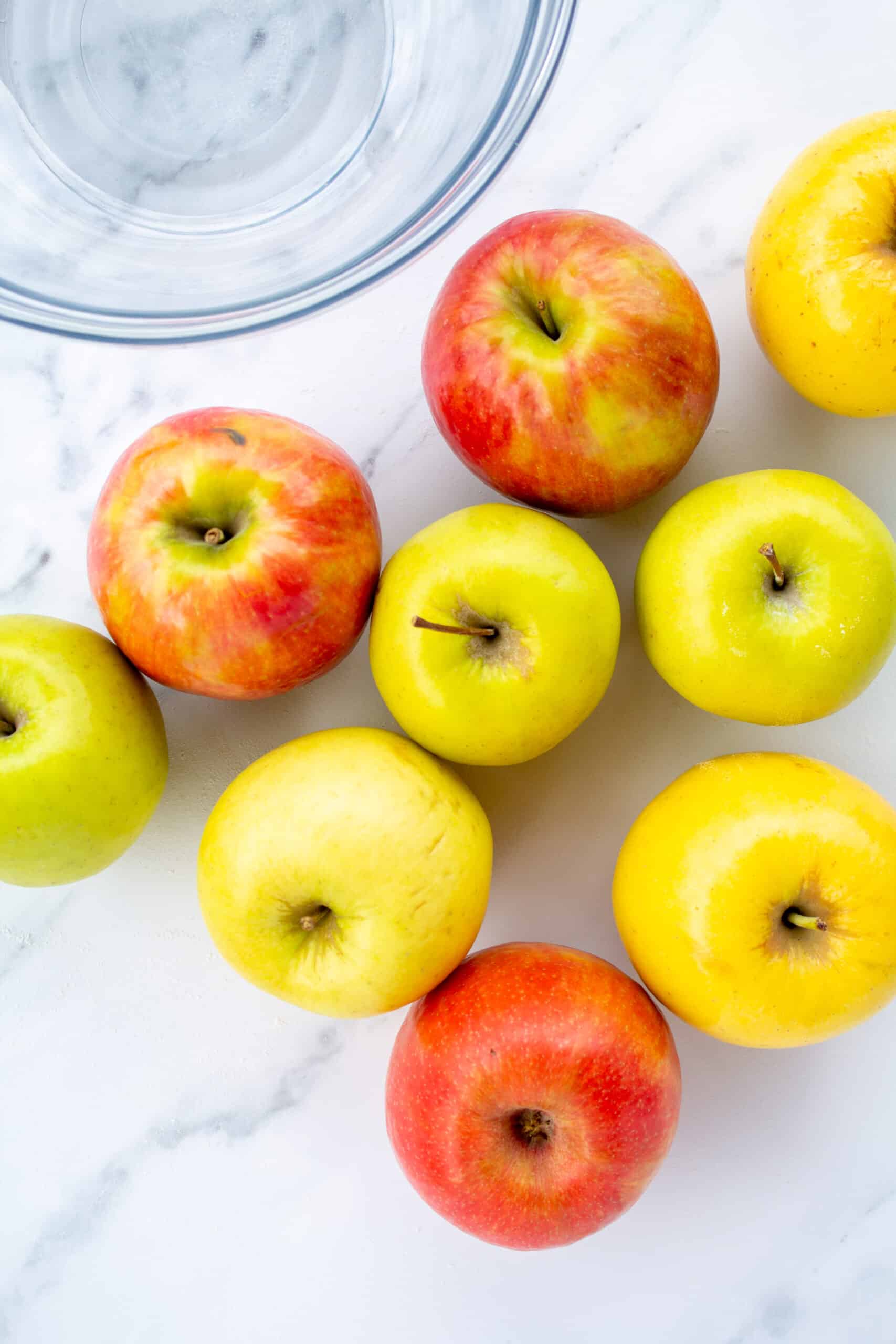 red and yellow apples spread out on a marble counter next to a glass bowl