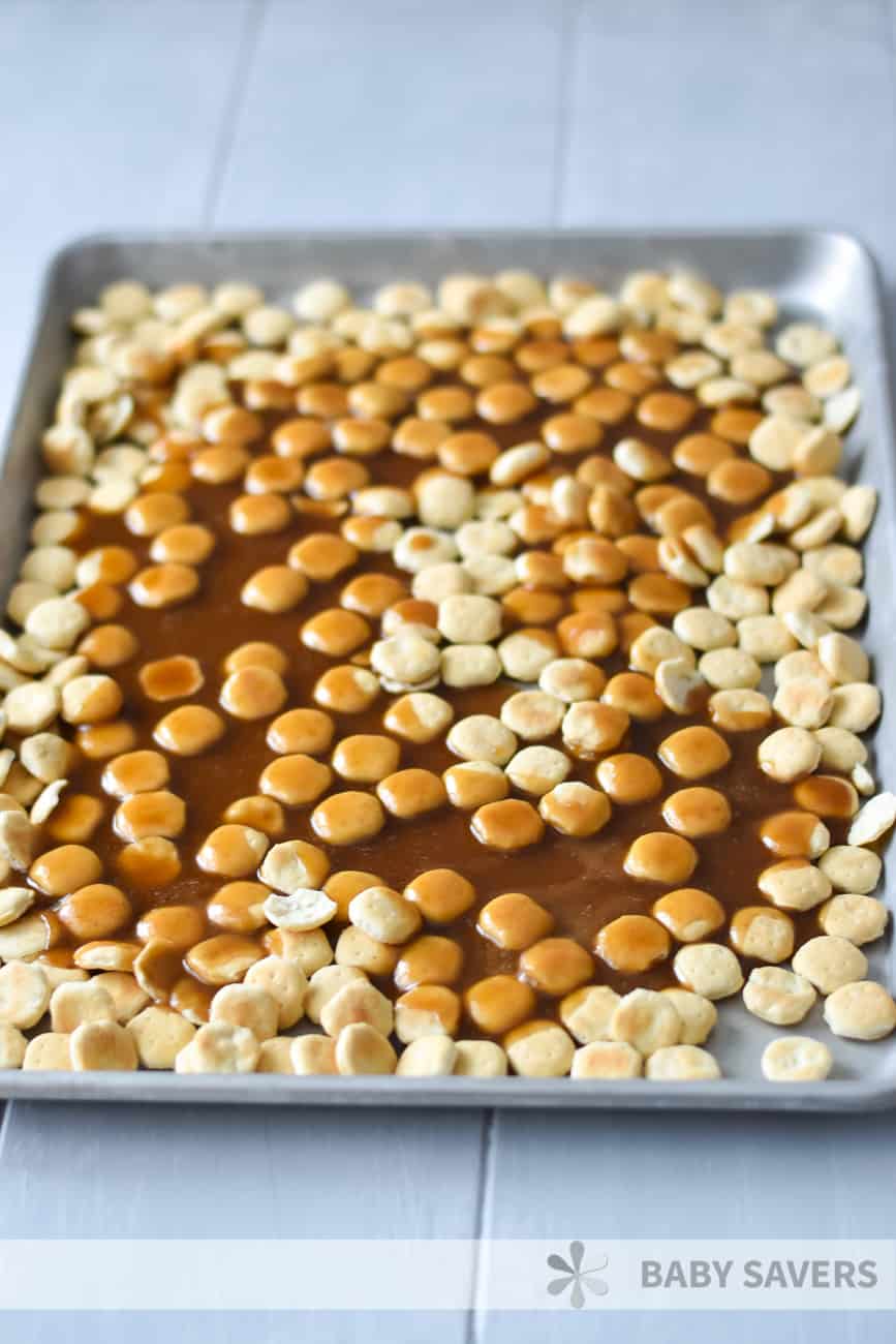 Oyster crackers spread on a baking sheet with caramel syrup poured over some of them, in the process of making oyster cracker snack mix 