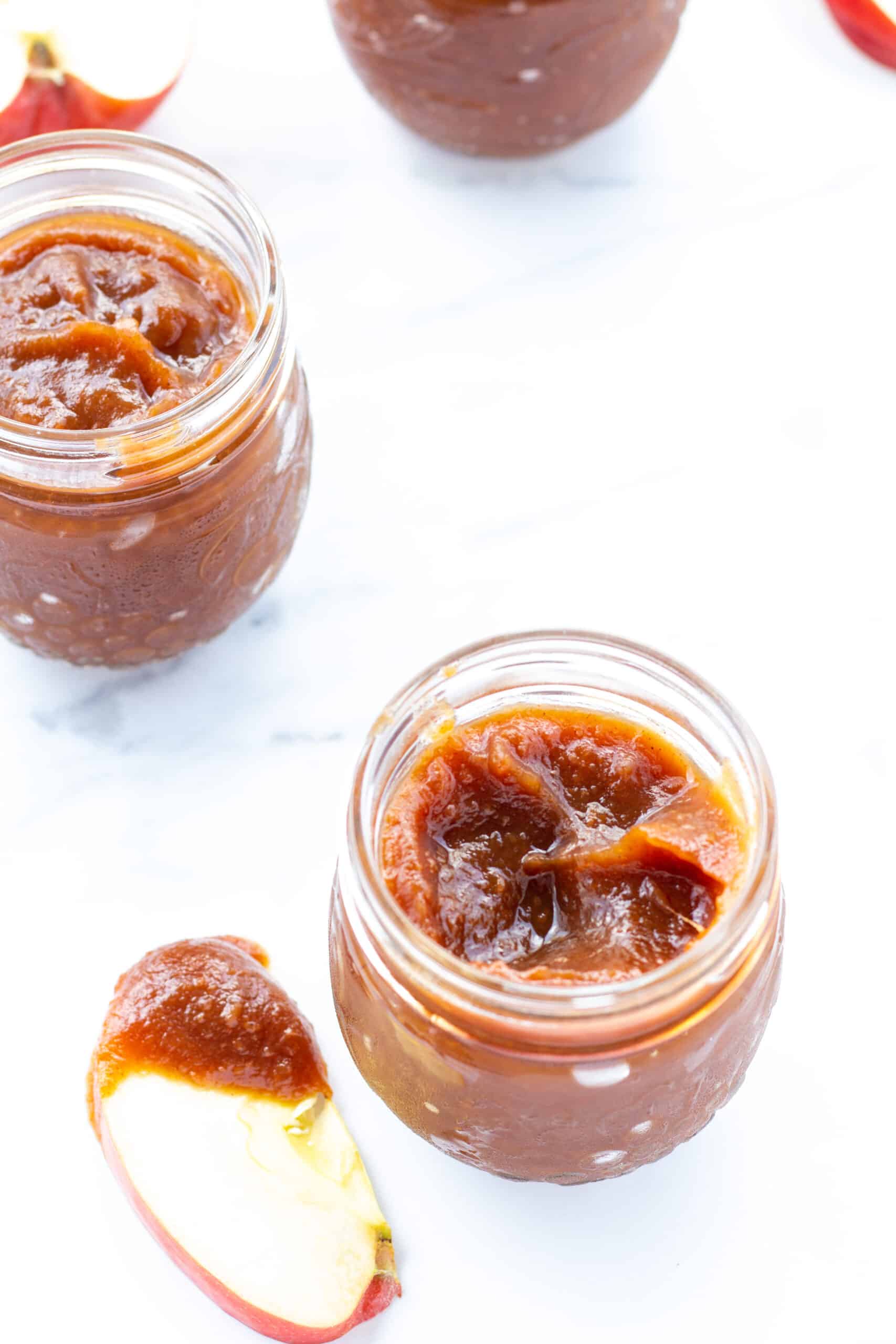 two jars of apple butter and one apple slice dipped in apple butter on a white counter