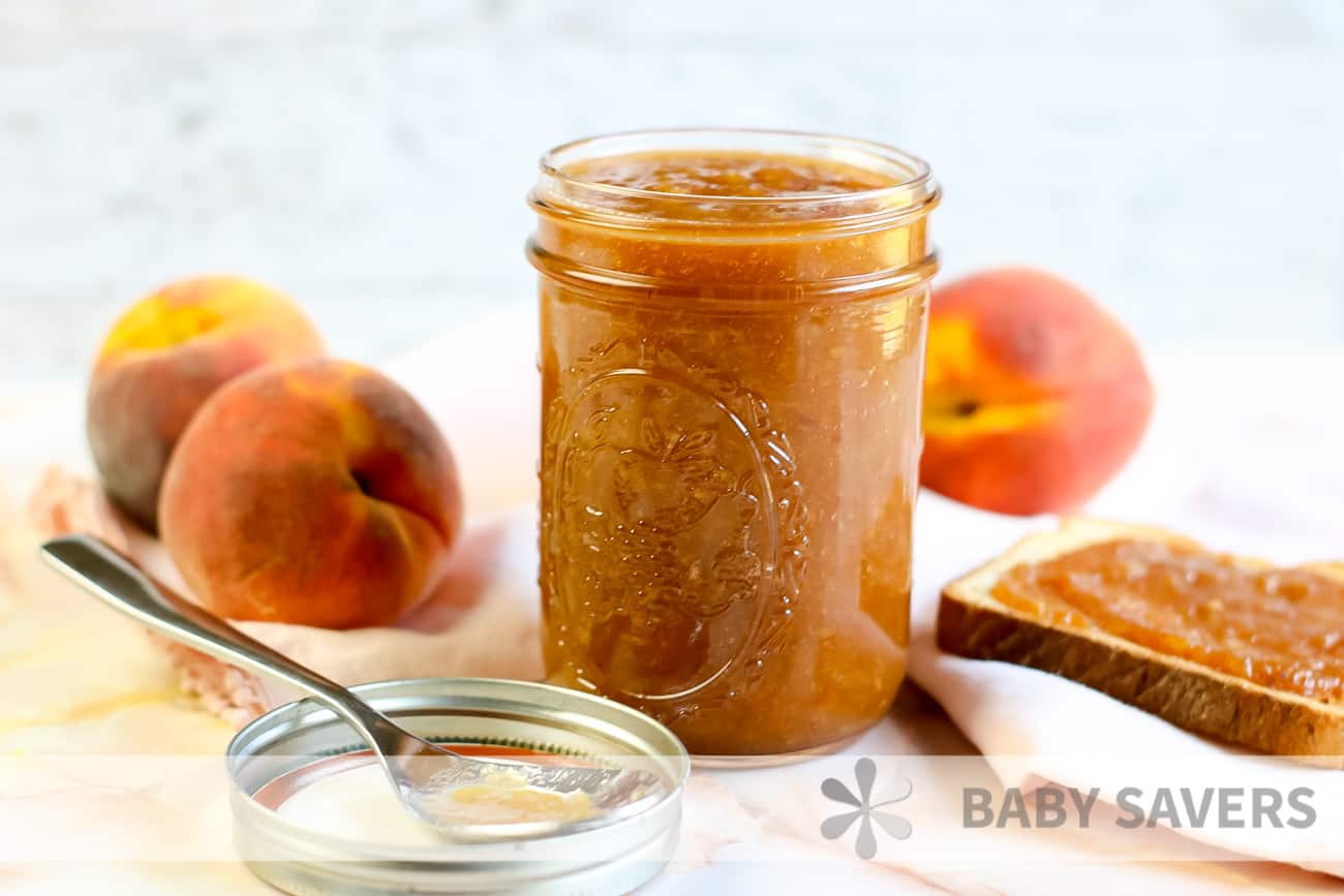 spoon with peach butter on it in front of a glass jar filled with peach preserves