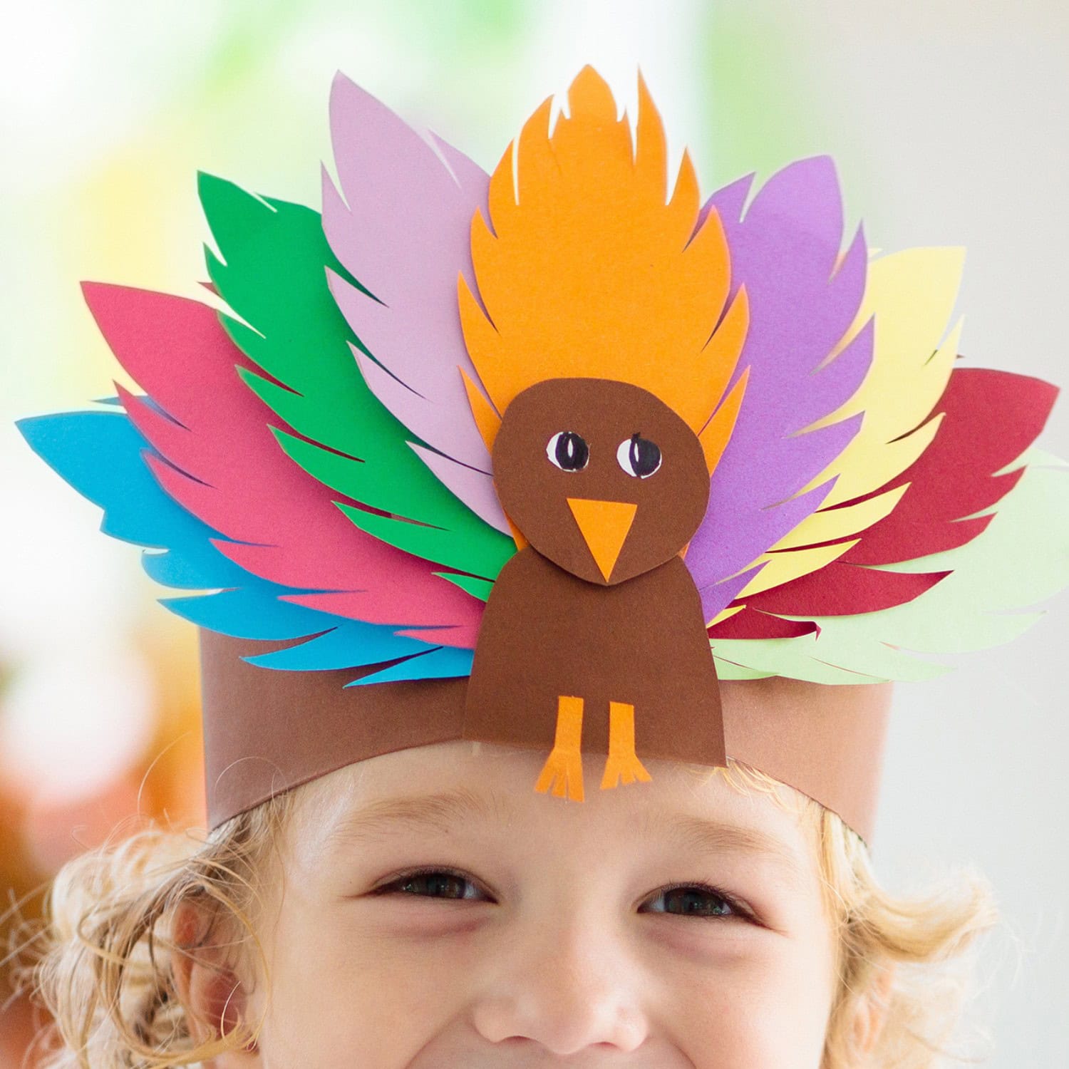 A toddler or preschool age young child proudly wears a colorful paper turkey hat adorned with multicolored feathers, showcasing an easy Thanksgiving craft. The hat features a brown turkey with orange feet and beak, set against a bright and cheerful background.