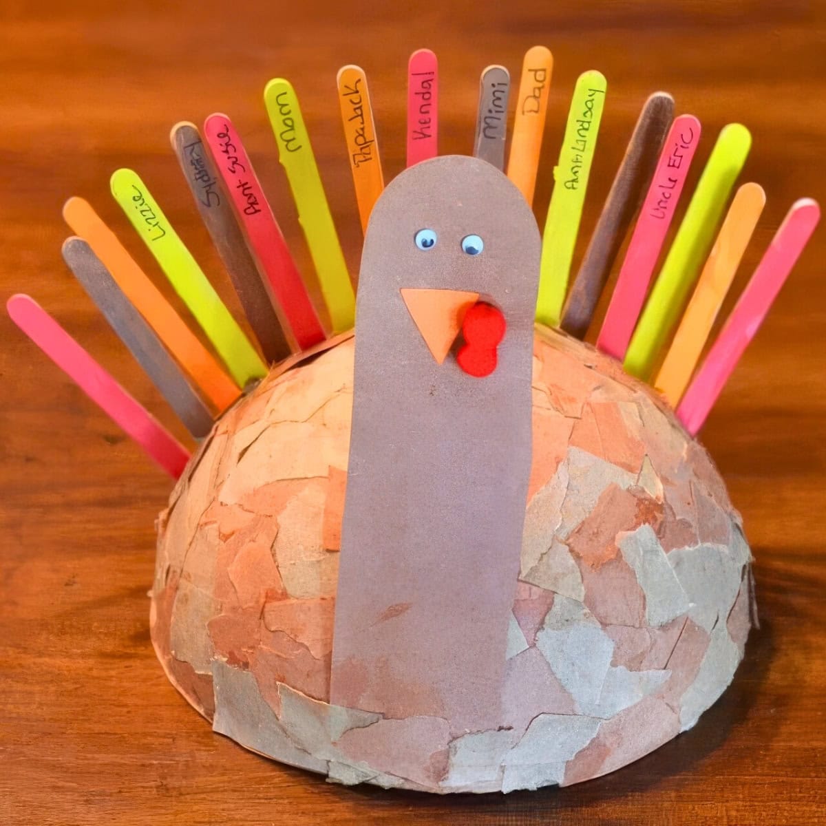 This easy Thanksgiving craft transforms a round base into a delightful turkey, adorned with brown paper pieces. Vibrant popsicle stick tail feathers proudly display names, while googly eyes and a red wattle complete the playful paper head.
