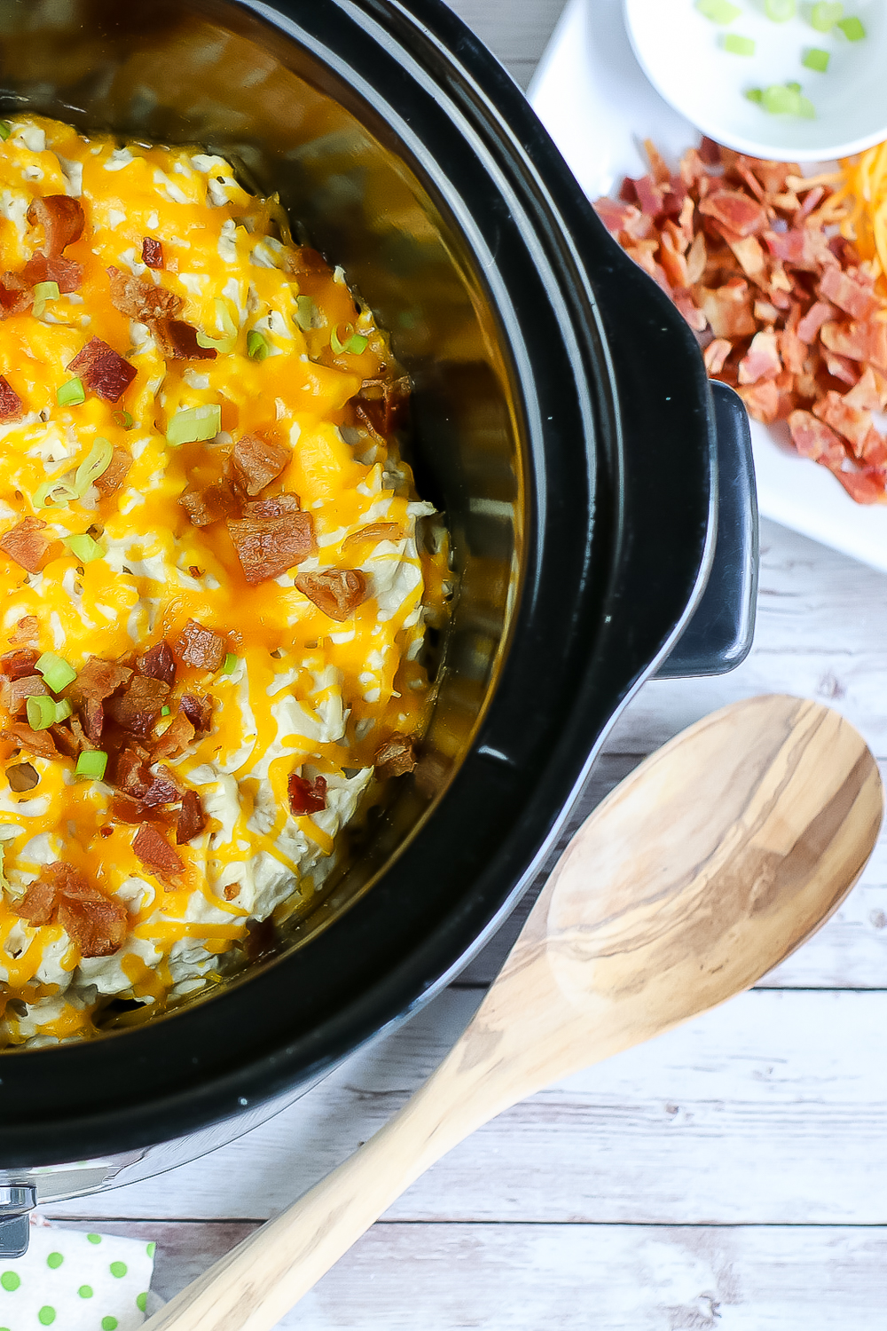 creamy crockpot chicken in crockpot with wooden spoon, sliced bacon, cheddar cheese and green onions