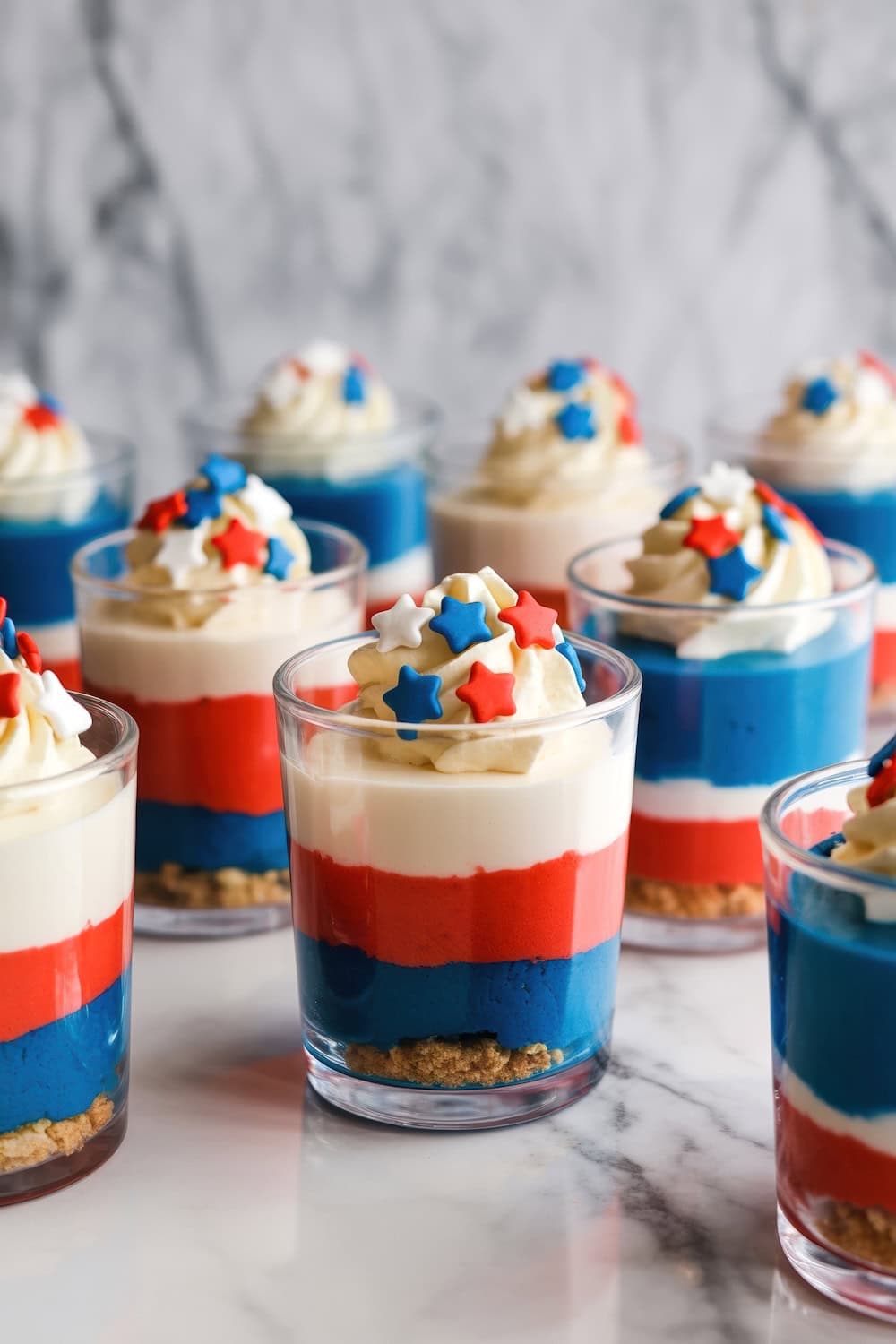 Several glass cups filled with layered desserts in blue, red, and white, topped with whipped cream and star-shaped sprinkles, make these a festive addition to your collection of Fourth of July recipes. Arranged on a marble surface for a patriotic touch.