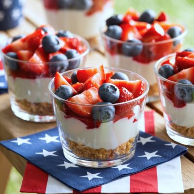 Four clear cups filled with layered cheesecake, strawberries, and blueberries sit on a wooden table, each on a patriotic star-spangled napkin—perfect for festive outdoor gatherings and ideal as Fourth of July recipes.