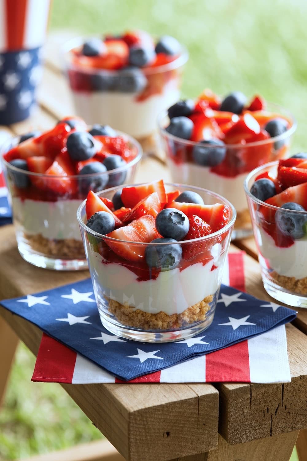 Four clear cups filled with layered cheesecake, strawberries, and blueberries sit on a wooden table, each on a patriotic star-spangled napkin—perfect for festive outdoor gatherings and ideal as Fourth of July recipes.