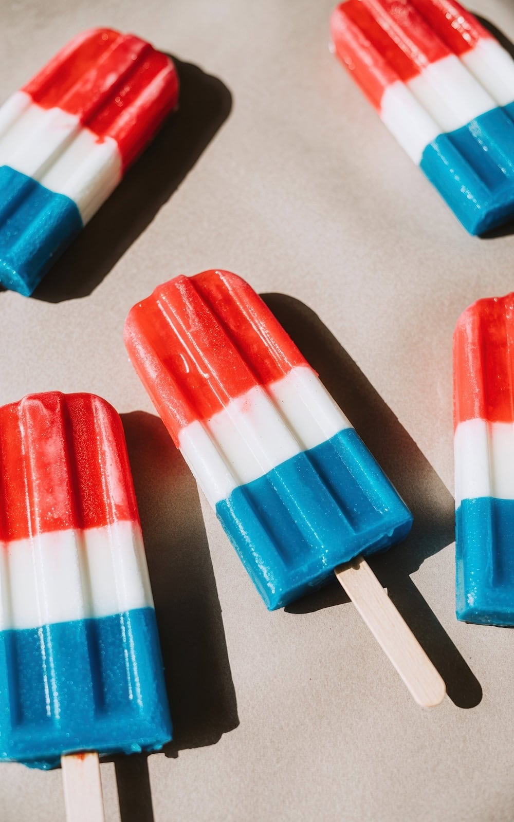 Five red, white, and blue popsicles are arranged on a light surface, sunlight casting shadows—a festive treat perfect for Fourth of July recipes, with horizontal stripes of red, white, and blue.
