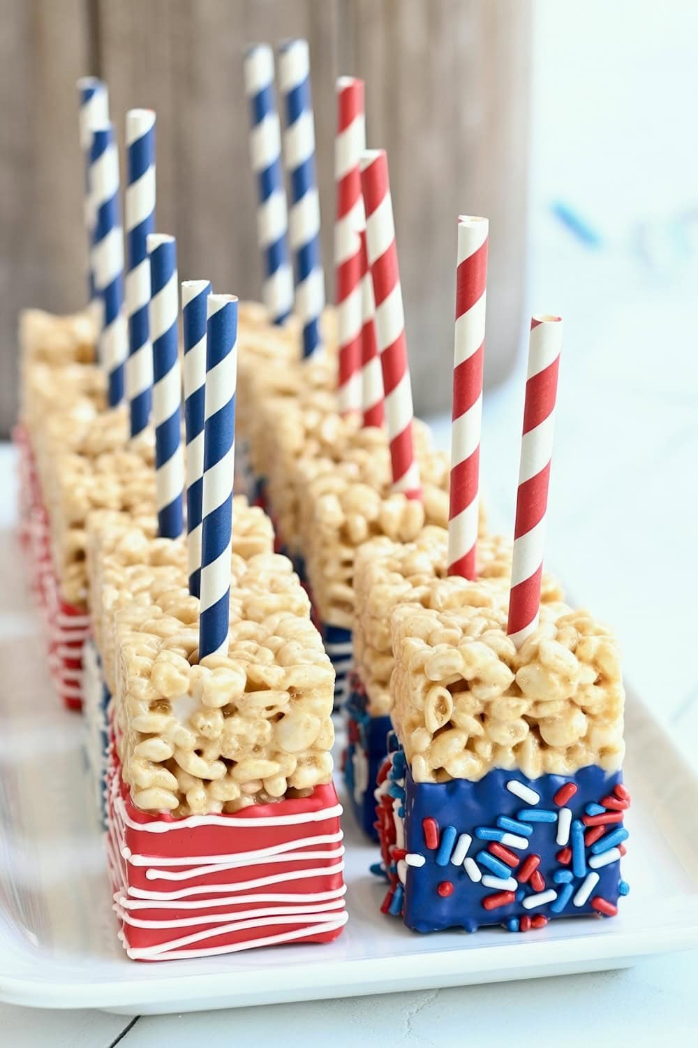 Rectangular Rice Krispies treats on a tray, each on a red or blue striped paper straw, with one end dipped in blue chocolate and decorated with red, white, and blue sprinkles—perfect for fun Fourth of July recipes.