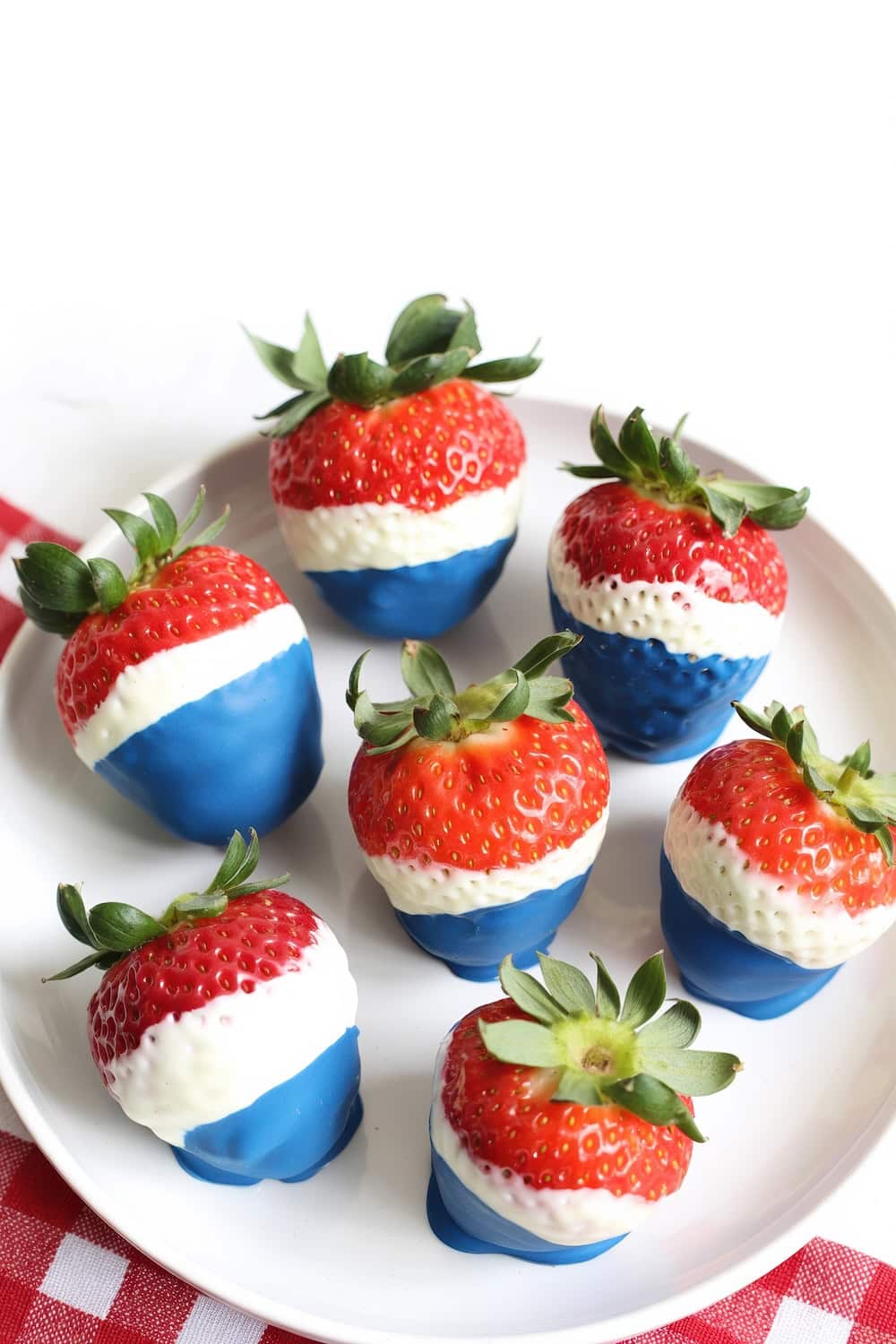A plate of strawberries dipped in white chocolate and blue candy coating creates a red, white, and blue layered effect—perfect for Fourth of July recipes. The plate sits on a red and white checkered cloth.