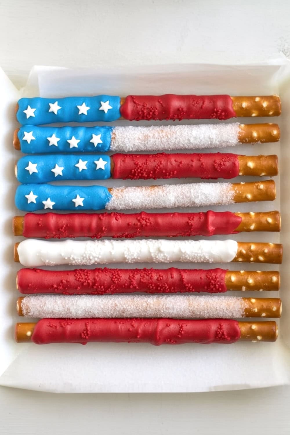 Pretzel rods decorated to resemble the American flag, with blue and white stars on the left and alternating red and white stripes. Perfect for Fourth of July recipes, these festive treats are arranged on parchment paper.