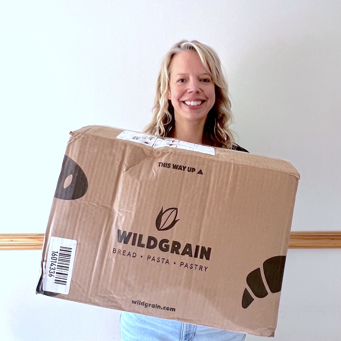 wildgrain reviews with a woman holding a wildgrain box shipped to her with a barcode and brand logo with bread, pasta and pastry with the URL of the website