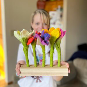 A person holds a wooden base with six colorful felt flowers, vibrant in hues of white, red, purple, yellow, and orange. The stems are green, set against a blurred interior with a fridge. Inspired by Lovevery play kit, this display of creativity is truly worth it.