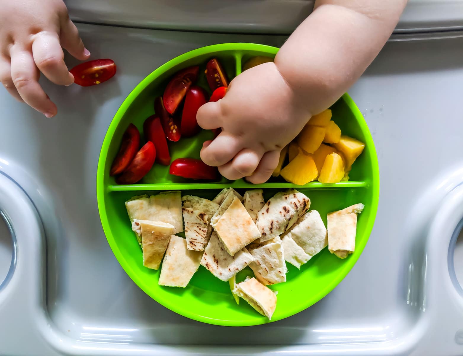 Toddler reaching for a colorful assortment of healthy breakfast options on a bright green divided plate.