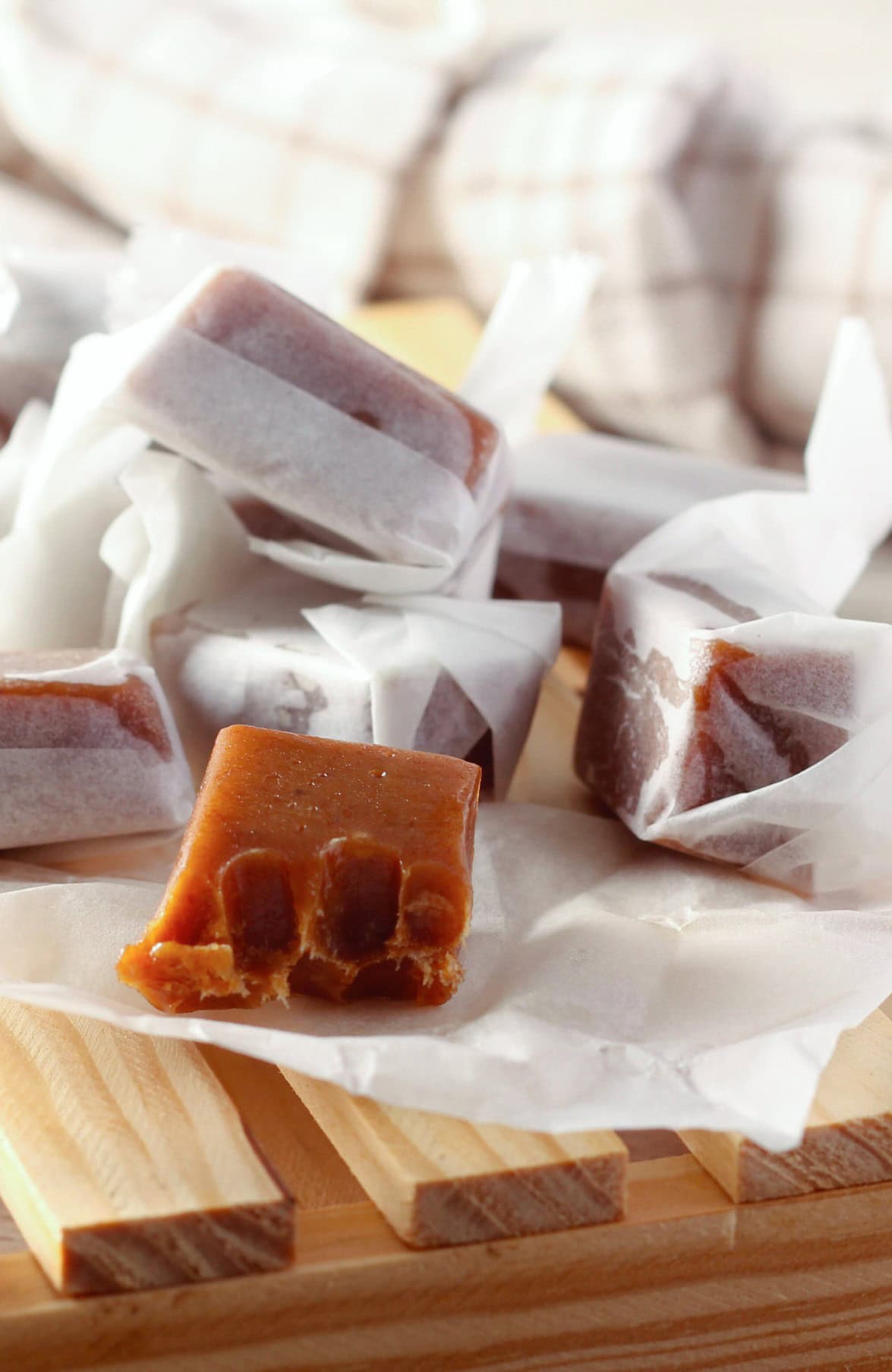 Homemade buttery dark brown sugar caramels wrapped in white paper, presented on a wooden tray with a checkered cloth in the background, on a wooden surface. The one in focus in the front is a closeup of an unwrapped caramel with a bite out of it.