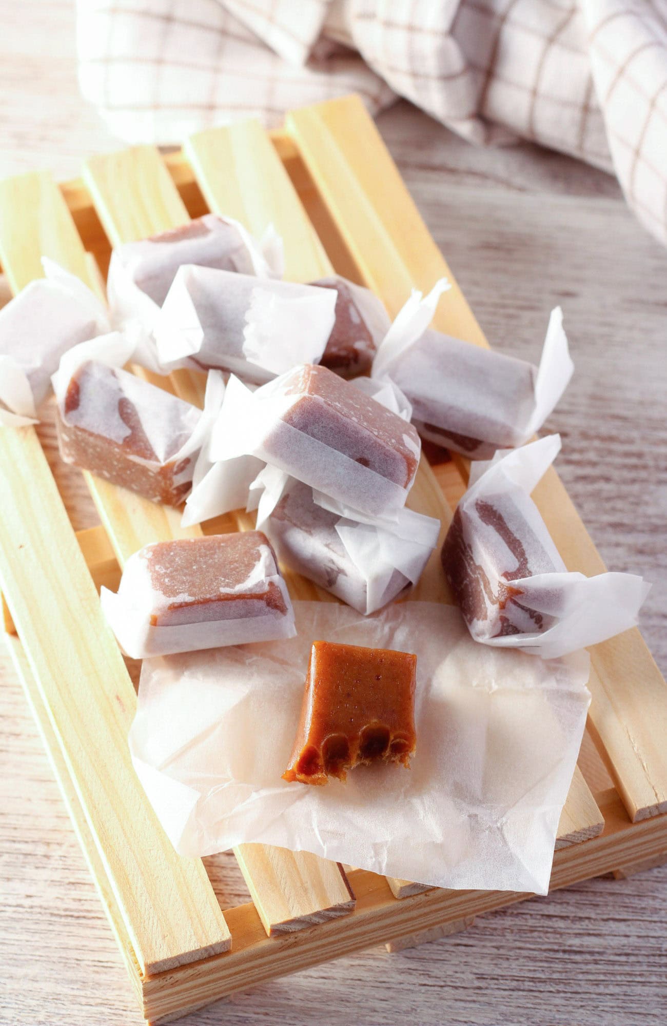 Homemade dark brown sugar caramels wrapped in white paper, presented on a wooden tray with a checkered cloth in the background, on a wooden surface. The one in focus in the front is a homemade dark brown sugar caramel candy with a bite out of it.