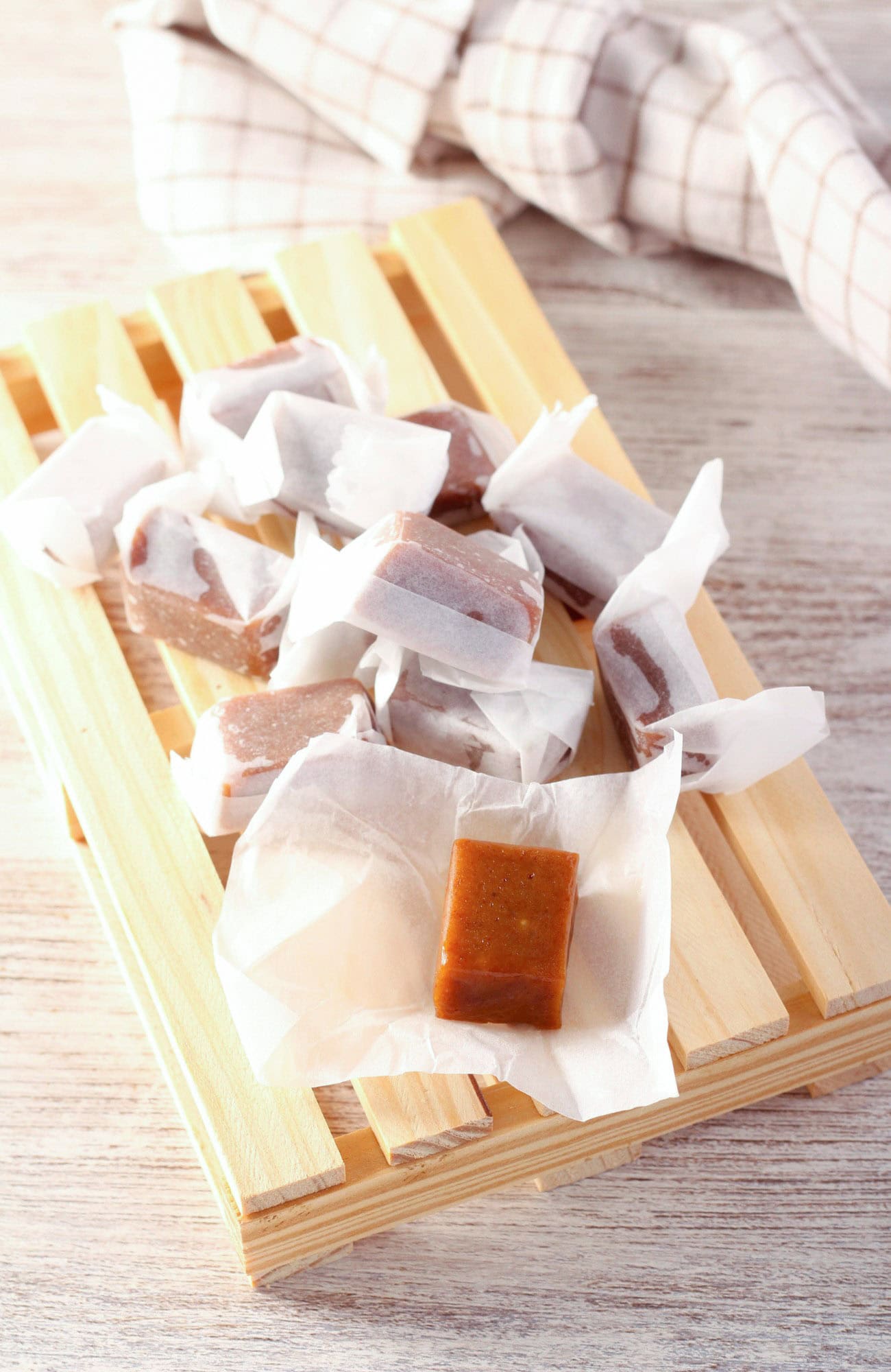 Homemade buttery dark brown sugar caramels wrapped in white paper, presented on a wooden tray with a checkered cloth in the background, on a wooden surface. There's an unwrapped caramel in focus at the front of the image.