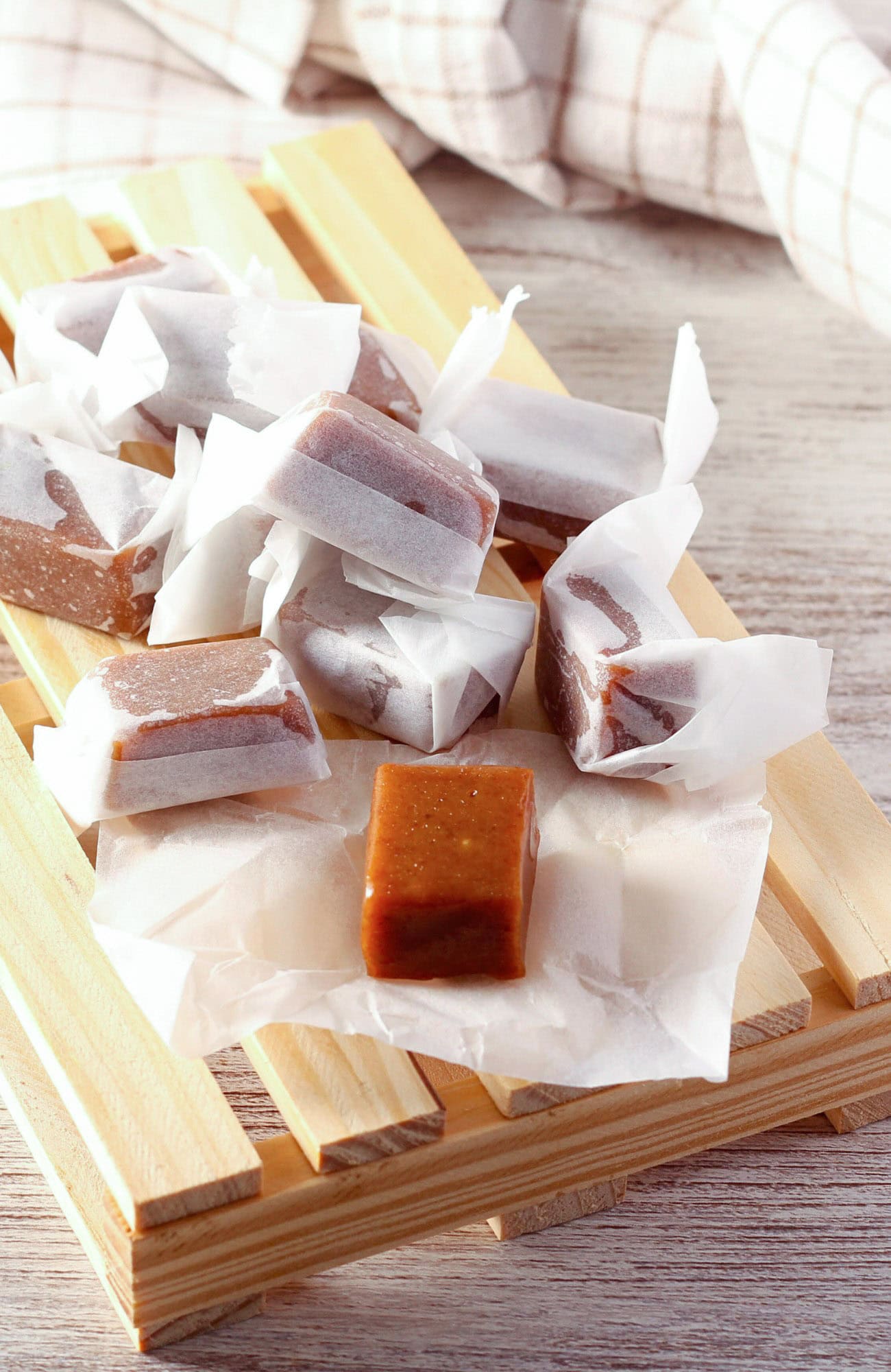 Homemade buttery dark brown sugar caramels wrapped in white paper, presented on a wooden tray with a checkered cloth in the background, on a wooden surface. 