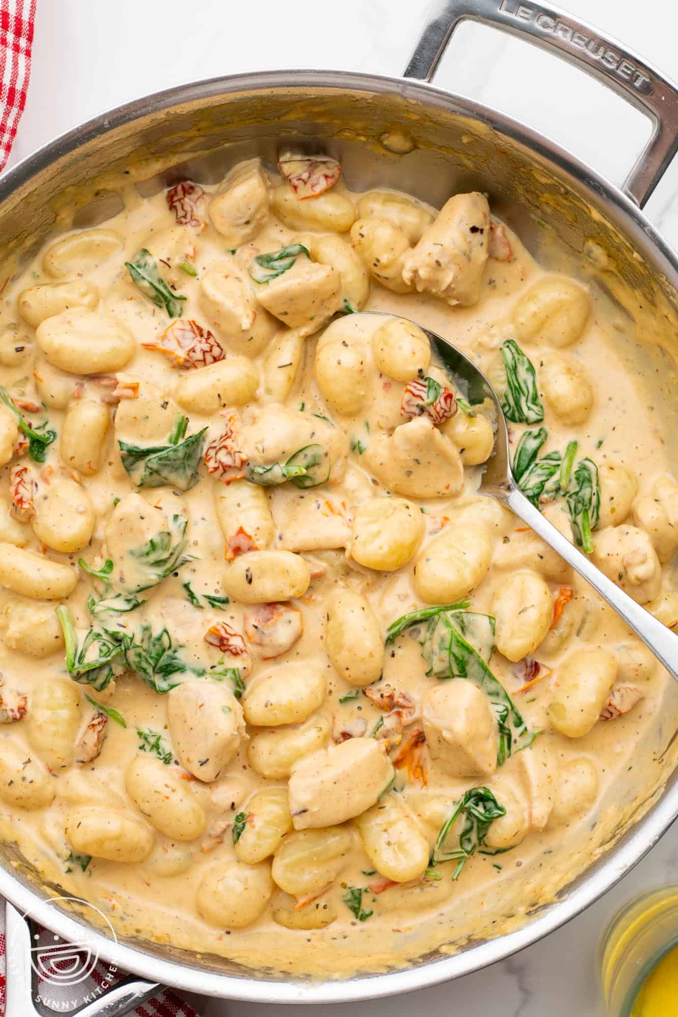A skillet filled with creamy gnocchi and sun-dried tomatoes garnished with fresh basil, displayed on a kitchen counter—perfect for busy families. A spoon rests inside the skillet.