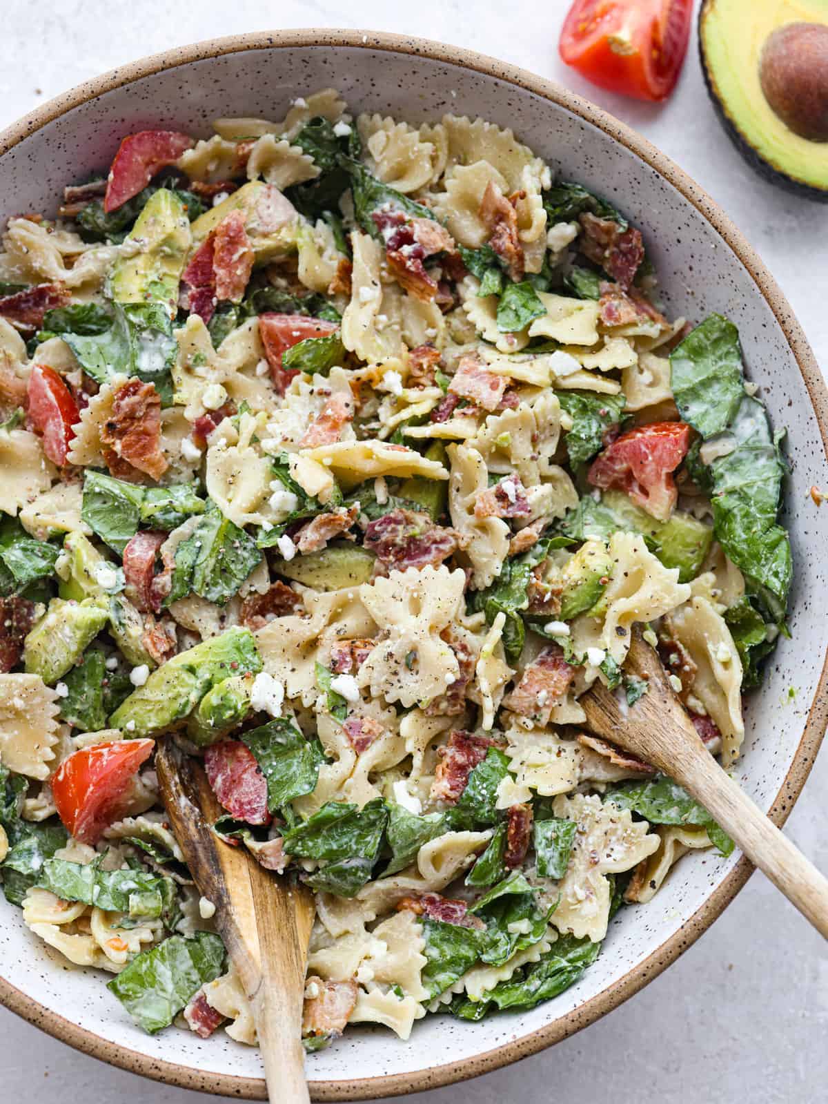 A bowl of pasta salad with bowtie noodles, spinach, sundried tomatoes, and asparagus, tossed with parmesan cheese and a wooden serving spoon—perfect for busy families.