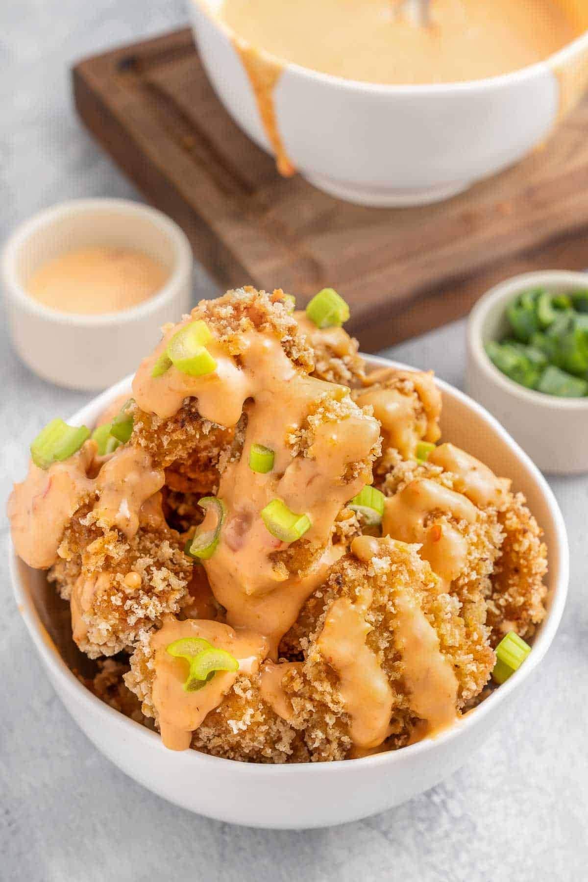 A bowl of crispy fried chicken pieces drizzled with creamy orange sauce, garnished with green onions, served on a wooden board with extra sauce on the side—perfect for busy families.
