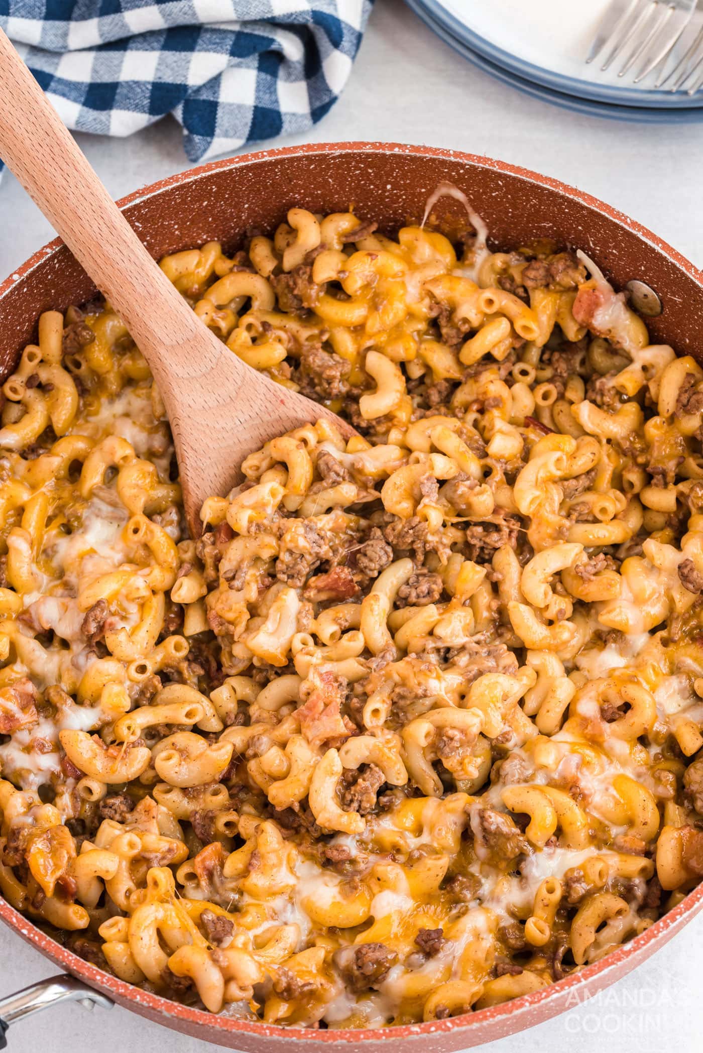 A skillet with cheesy beef and macaroni, perfect for busy families, with a wooden spoon stirring the mixture, framed by a blue and white checkered napkin and a plate in the background.