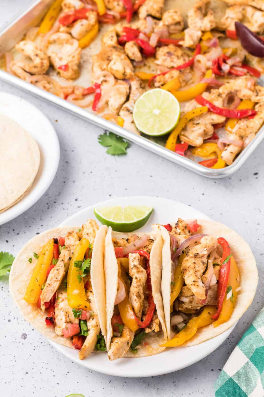 Three chicken fajitas with colorful bell peppers and onions in soft tortillas, garnished with lime wedges and cilantro, alongside a tray of ingredients—perfect for busy families.