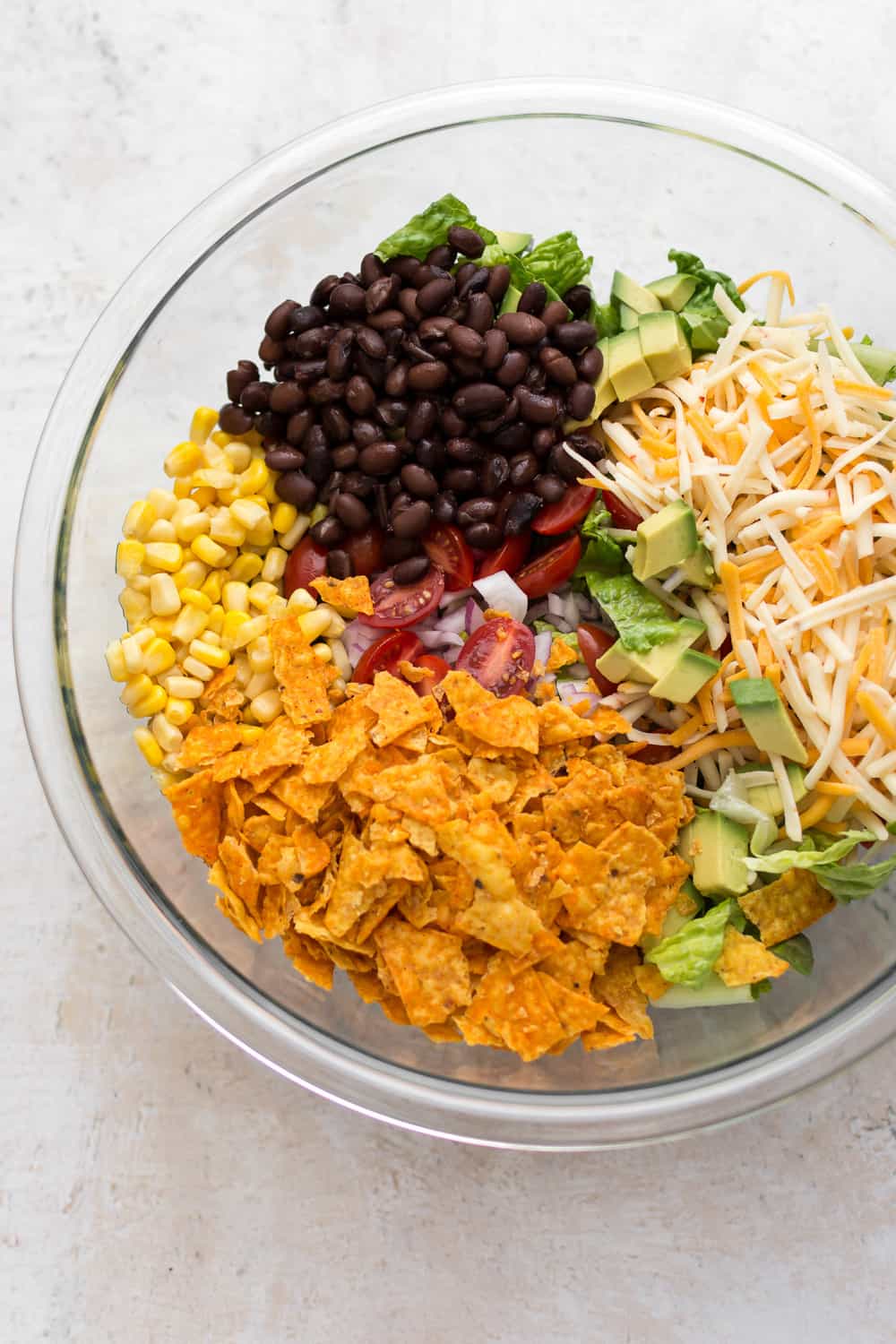 A colorful salad in a glass bowl with sections of black beans, chopped tomatoes, corn, crushed tortilla chips, diced avocado, shredded cheese, and lettuce on a white surface makes for easy dinner ideas
