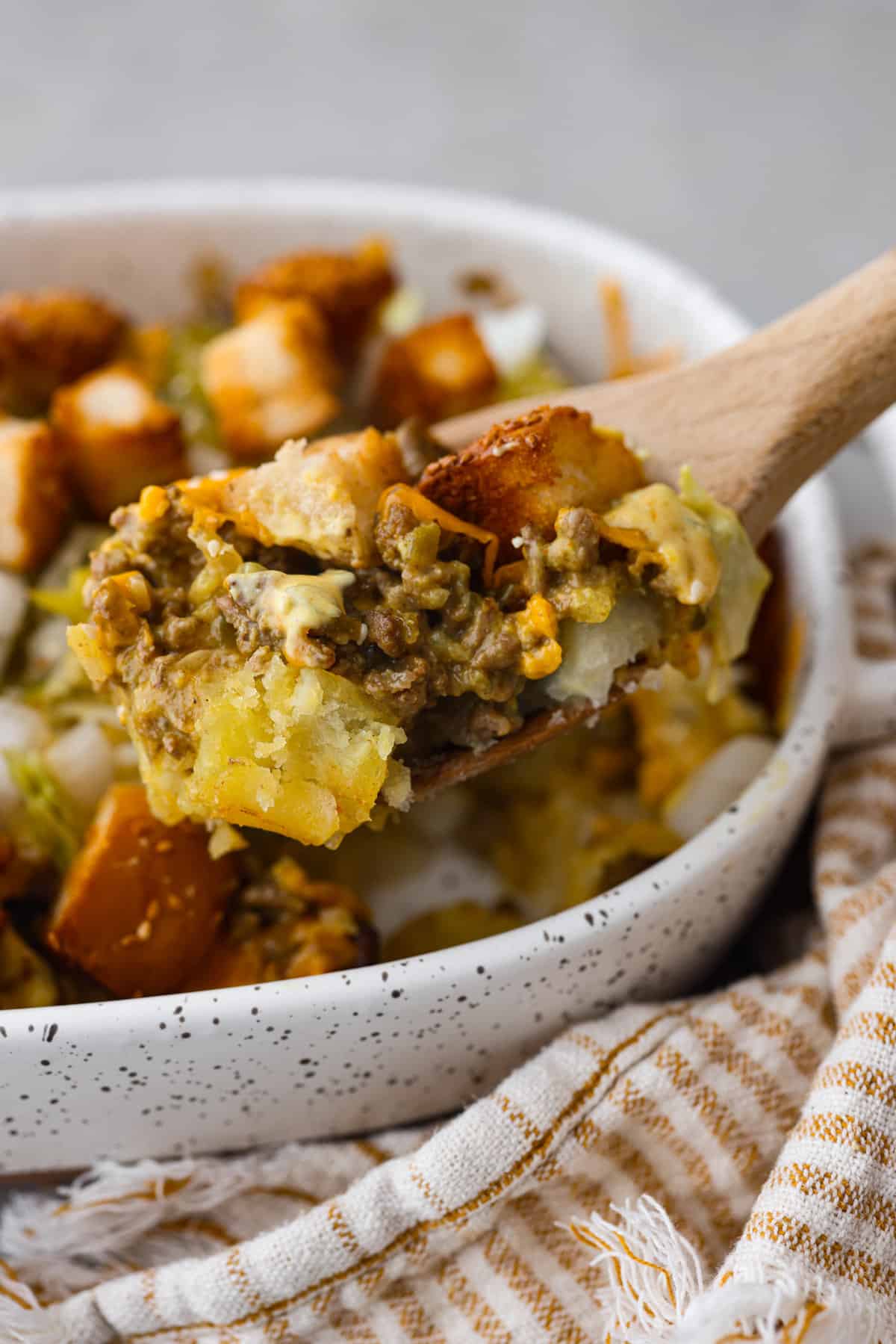 A spoon lifts a portion of shepherd's pie from a speckled bowl, showing layers of minced meat and vegetables topped with golden-brown mashed potatoes, making it an easy dinner idea for busy families