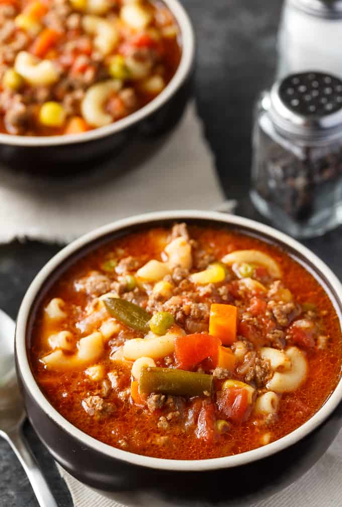 A bowl of hearty vegetable and ground meat soup with corn, green beans, and macaroni, served on a dark tabletop accompanied by a pepper shaker; perfect for busy families.