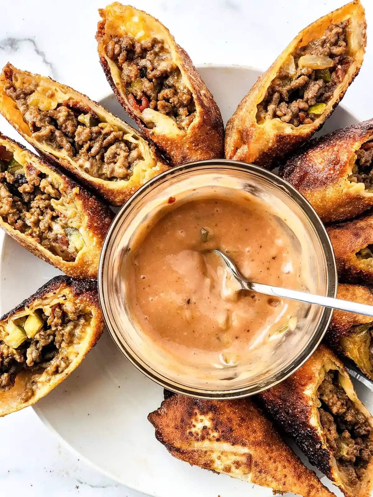 Plate of crispy egg rolls cut in half, filled with meat and vegetables, served with a creamy dipping sauce in a glass bowl on a white marble surface, perfect for busy families.