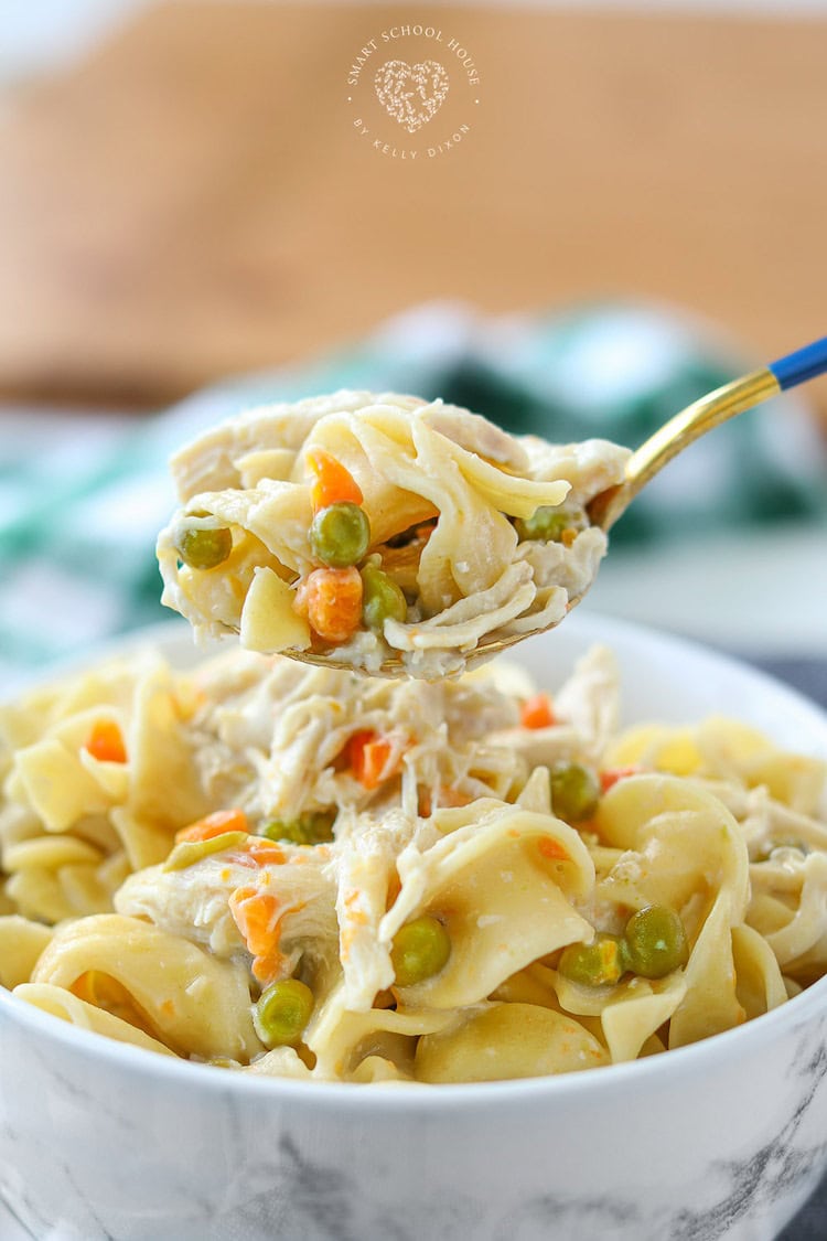 A close-up of a fork lifting creamy chicken noodle soup from a white bowl, featuring chunks of chicken, peas, and carrots, with a soft-focused background. This meal is perfect for busy families