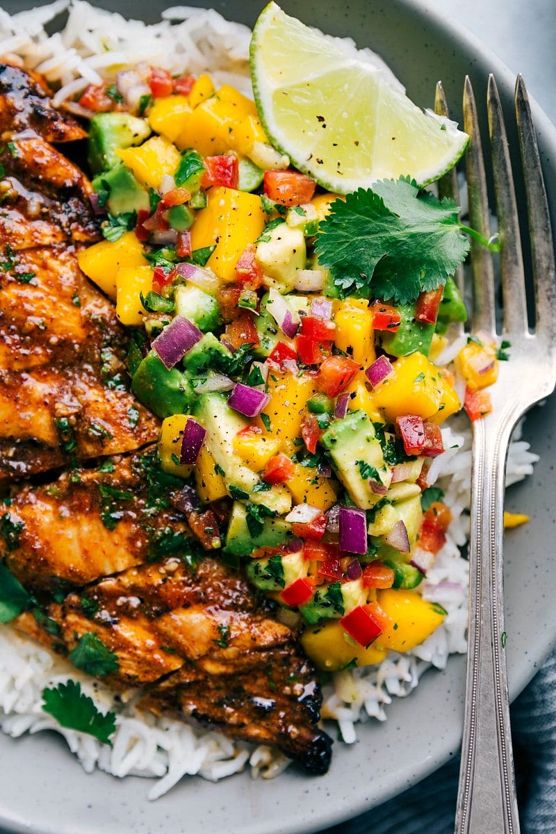 Grilled chicken breast topped with mango salsa, served on a bed of rice garnished with lime and cilantro, accompanied by a fork on a patterned plate. Perfect for busy families.